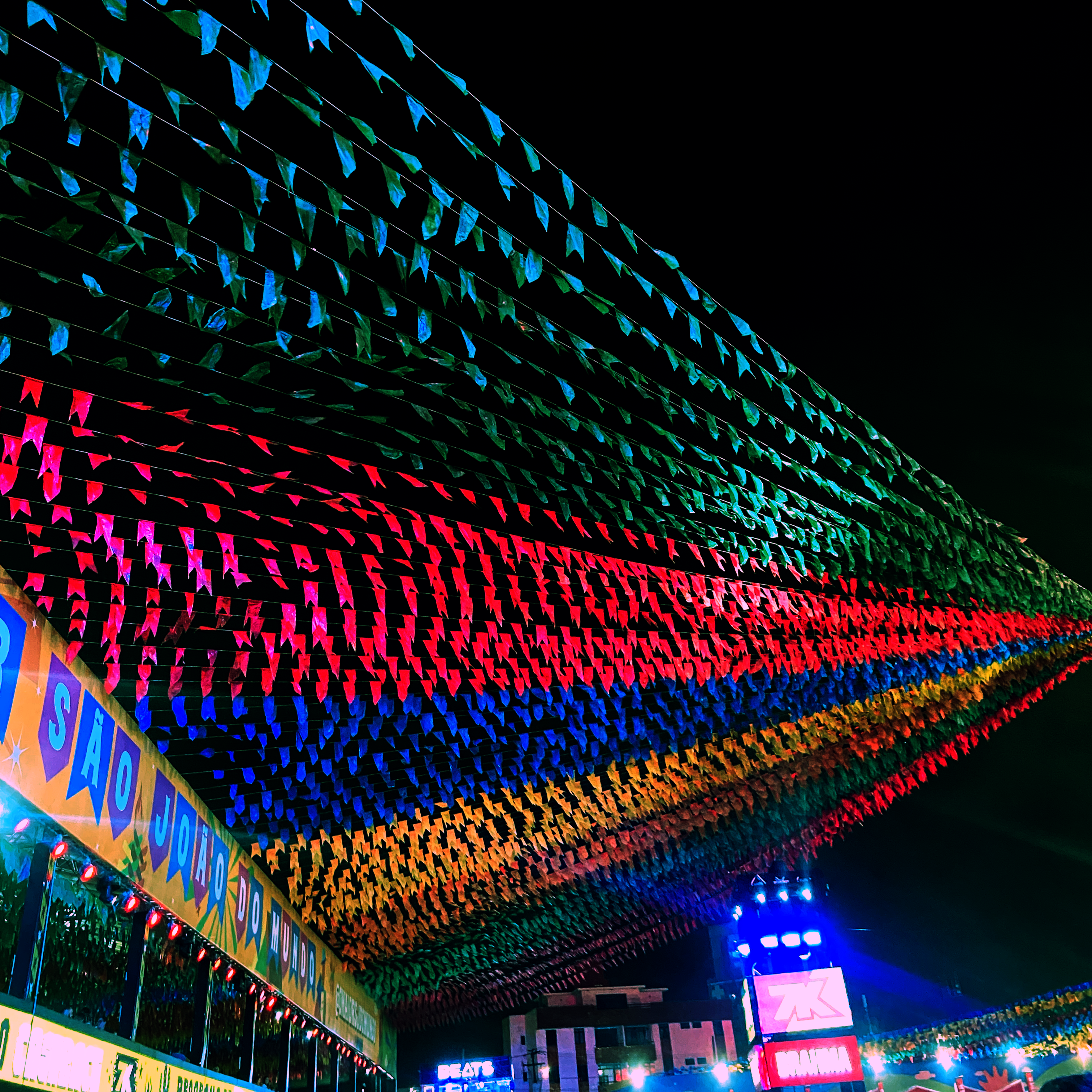 Céu escuro coberto por uma decoração de festa junina, com fileiras de bandeirinhas em cores vibrantes como verde, azul, vermelho e amarelo, iluminadas à noite.